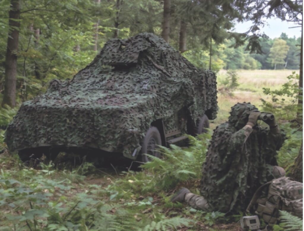 Camouflaged military vehicle and soldier concealed in woodland terrain.